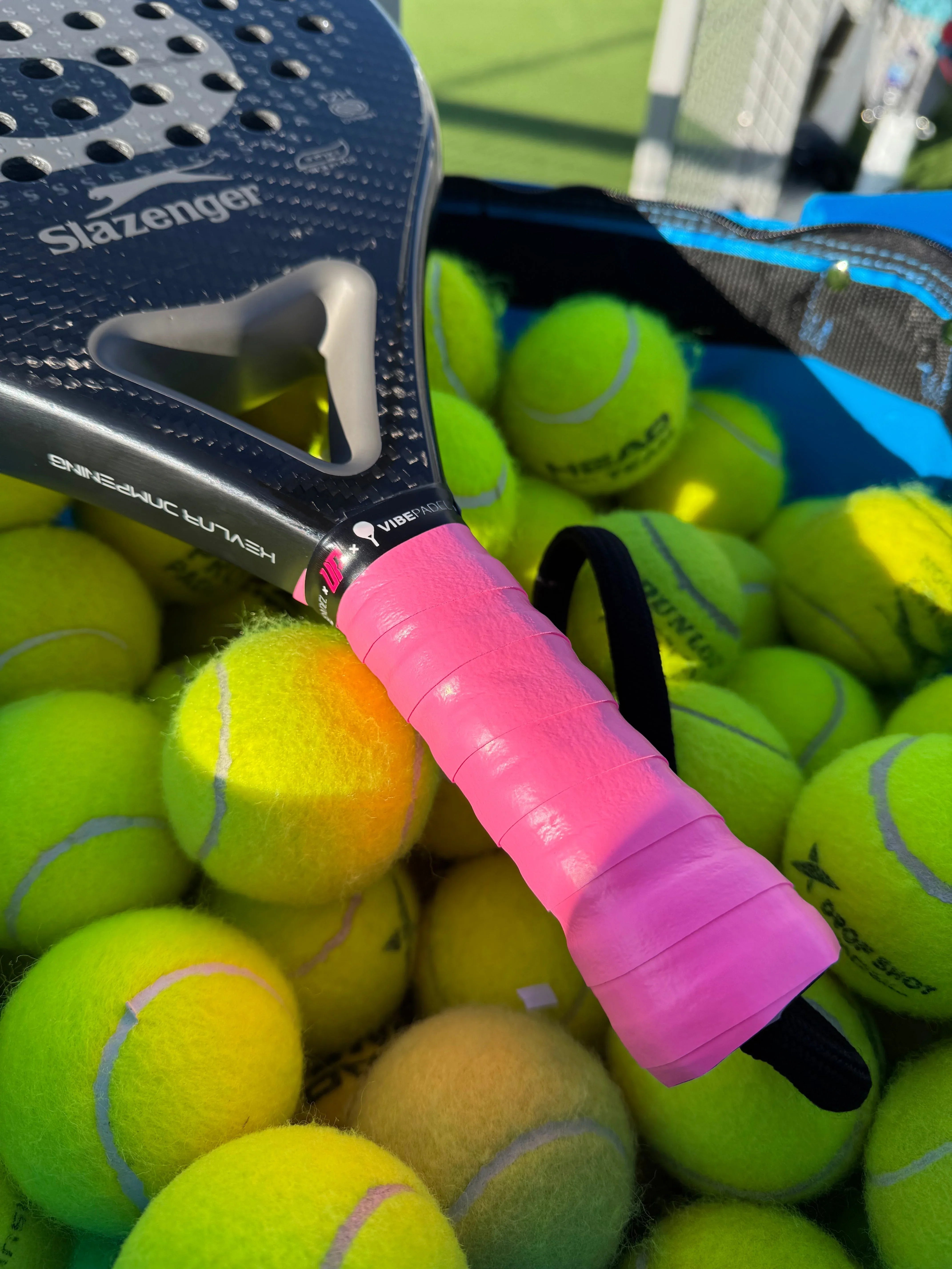 Slazenger padel racket with pink grip lying on a pile of yellow tennis balls on outdoor court