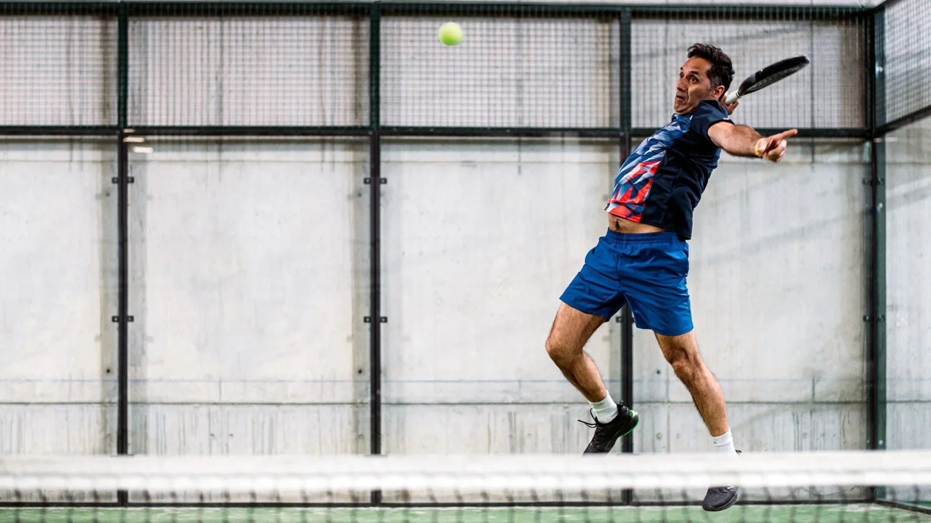 Man in blue sportswear jumping to hit a ball with a racket on an indoor padel court