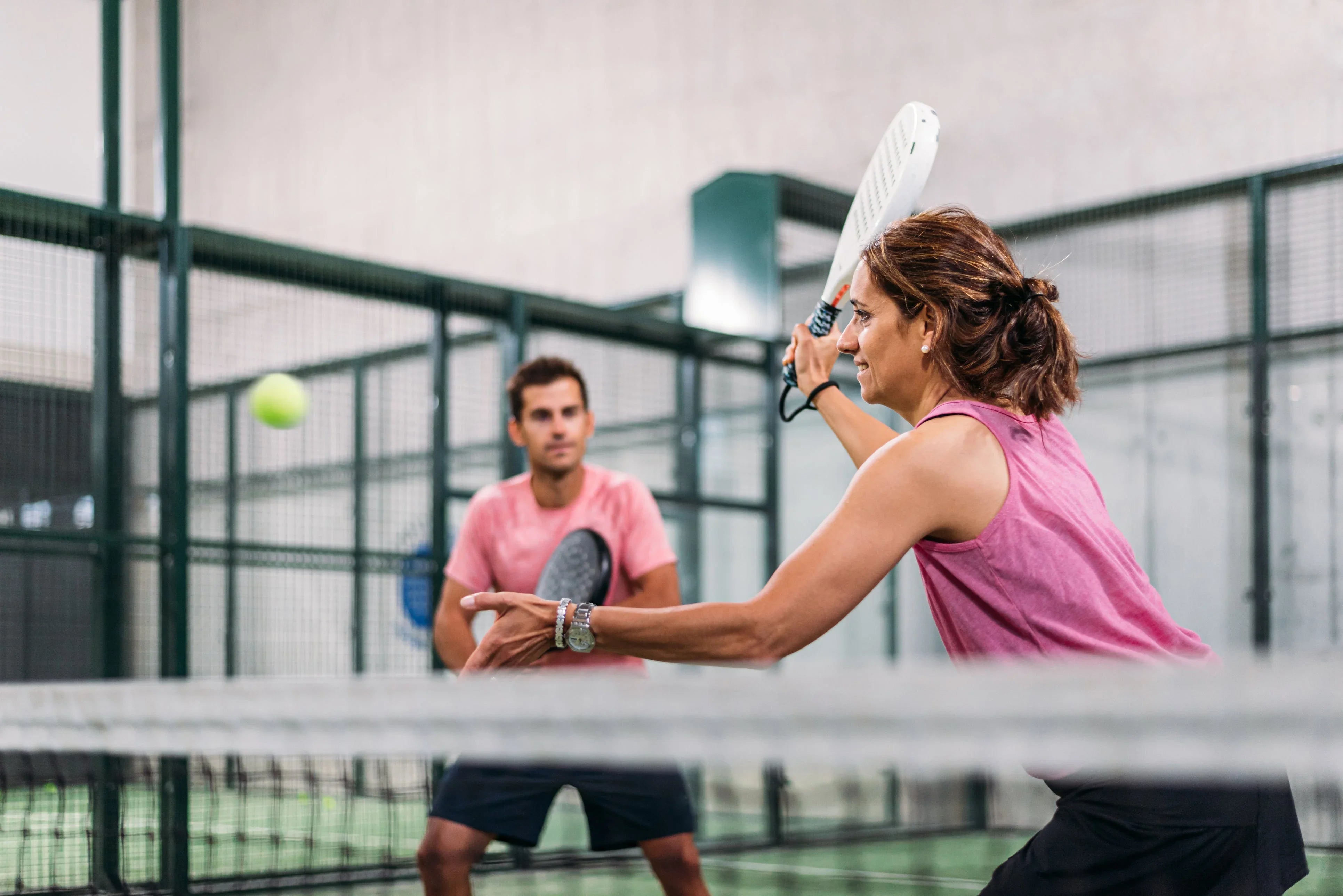 Two people playing padel indoors, woman in pink preparing to hit ball with racket