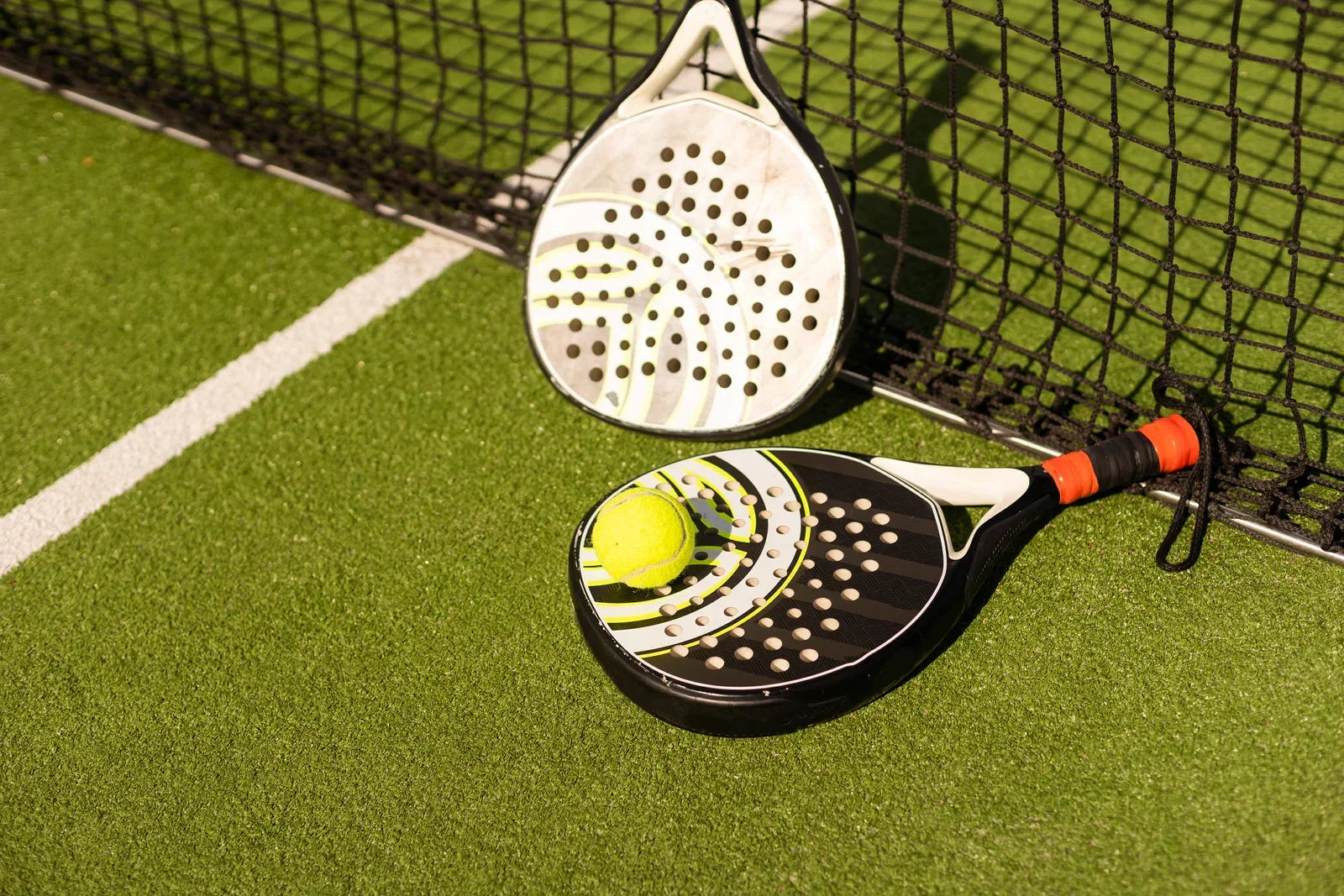 Two modern padel rackets and a yellow ball on an artificial grass padel court near the net